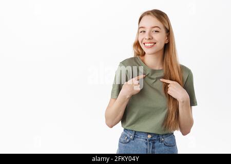 Ich bin es. Lächelnde, nette junge Frau, die mit schüchternem Gesichtsausdruck auf sich zeigt, sich selbst fördert, über sie spricht, prahlend, über Weiß stehend Stockfoto