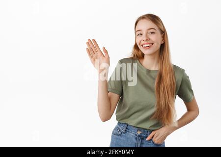 Freundliches blondes Mädchen winkt Hand zu sagen, Hallo. Junge Frau lächelt und grüßt, begrüßt Sie, begrüßt Menschen, steht in Sommerkleidung dagegen Stockfoto