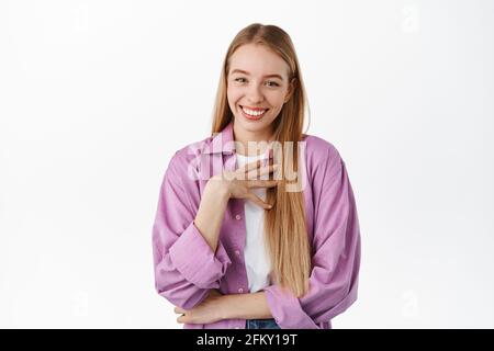 Bescheidenes, nettes blondes Mädchen, das berührt und geschmeichelt aussieht, erfreut lächelt, für etwas dankt, im stilvollen violetten Hemd gegen Weiß stehend Stockfoto