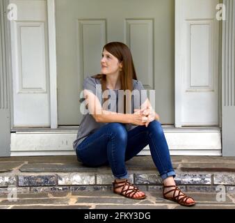 Die schöne junge Frau sitzt vor ihrem Haus auf der vorderen Treppe. Sie schaut weg, verloren in Gedanken. Sie hat Jeans und ein gestreiftes Hemd. Stockfoto