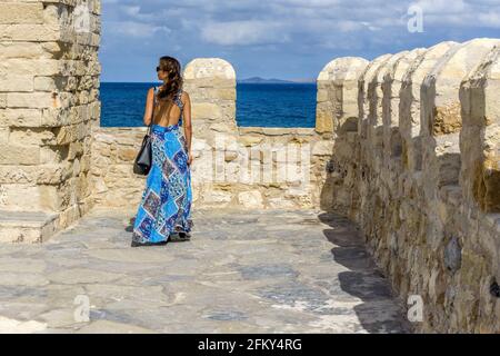 Heraklion, Kreta, Griechenland. Eine junge Frau in blauem Kleid auf dem Dach der Festung Koules bewundert den Blick auf das Meer. Sonniger Tag, wolkenblauer Himmel Stockfoto