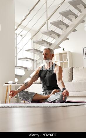 Gesunder älterer tätowierter Mann mittleren Alters, der zu Hause in Yoga-Pose meditiert. Stockfoto