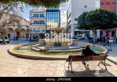 Heraklion, Kreta, Griechenland. Morosini-Brunnen (der sogenannte Löwenbrunnen) am Kallergen-Platz in der Stadt Heraklion. Der alte Mann sitzt auf einer Bank Stockfoto