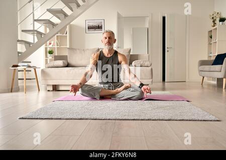 Ruhiger, gesunder, gelassener tätowierte Mann mittleren Alters, der zu Hause in Yoga-Pose meditiert. Stockfoto