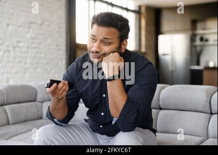 Gelangweilter junger inder, der auf der Couch sitzt und Fernsehen schaut, Mischling, der die Kanäle wechselt, fühlt Traurigkeit und Einsamkeit, fauler Kerl verbringt das Wochenende mit tv-Shows Stockfoto