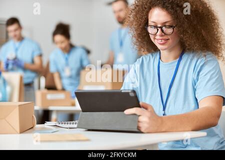 Junge Frau, Freiwillige in blauer Uniform mit Tablet-pc und lächelnd, während sie drinnen sitzt. Team-Sortierung, Verpacken von Artikeln im Hintergrund Stockfoto