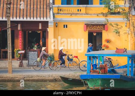 Hoi an, Vietnam - März 6 2019: Vietnamesische Radler entlang des Thu Bon River und lebendige, farbenfrohe, gelbe traditionelle Gebäude und Straßen von H Stockfoto