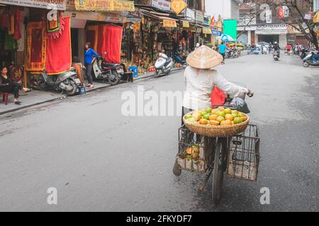 Hanoi, Vietnam - 28 2019. Februar: Ein lokaler vietnamesischer Straßenhändler verkauft in einem traditionellen konischen Non-la- oder Blumenhut Früchte von der Rückseite ihres Bi Stockfoto