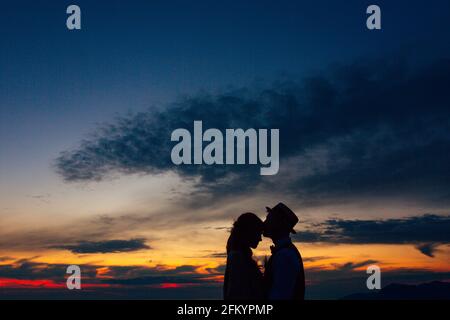 Silhouette des Bräutigams im Hut küsst Braut auf die Stirn bei Sonnenuntergang Stockfoto