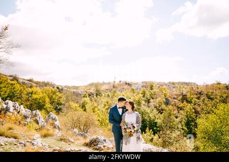 Der Bräutigam umarmt die Braut in einem karierten Schal mit einem Bouquet Von Blumen in ihren Händen auf dem Hintergrund von Felsen Und grünen Pflanzen Stockfoto