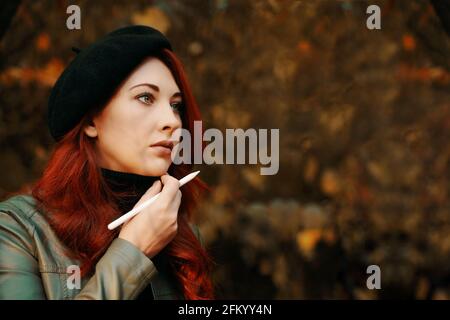 Brütende rothaarige Frau im Park. Schöne Frau in einer Baskenmütze und Lederjacke. Mann mit Stift, Designer, Freiberufler. Herbstporträt. Warten auf Inspiration. Stockfoto