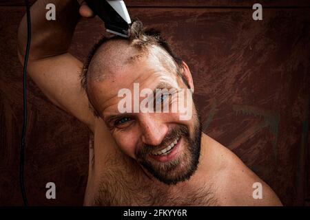 Ein erwachsener, fröhlicher Mann mit Bart rasiert sich zu Hause den Kopf. Der Typ schaut in den Badezimmerspiegel und benutzt den elektrischen Trimmer. Vorderansicht Stockfoto