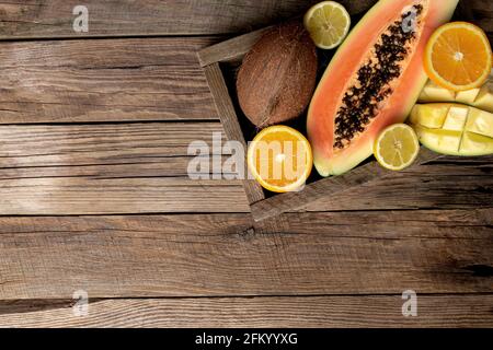 Frische tropische Früchte in einem Holzlieferkarton auf einem Holzhintergrund. Papaya, Orange, Kokosnuss, Mango und Zitrone Draufsicht flach Lay mit Kopierplatz für Stockfoto