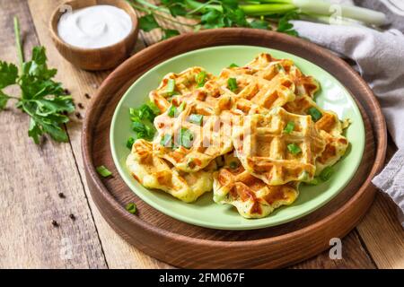 Nicht süße Waffeln mit Kartoffeln, Erbsen, Paprika, Käse werden mit Sauerrahm und Kräutern auf einem Teller auf einem rustikalen Tisch serviert. Stockfoto