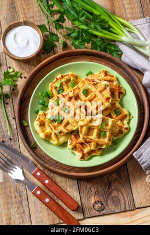 Nicht süße Waffeln mit Kartoffeln, Erbsen, Paprika, Käse werden mit Sauerrahm und Kräutern auf einem Teller auf einem rustikalen Tisch serviert. Stockfoto