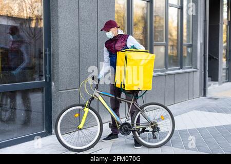 Junger Kurier in medizinischer Maske liefert Lebensmittel mit gelbem Thermalrucksack, fährt mit dem Fahrrad in der Stadt. Konzept für den Lieferservice von Lebensmitteln Stockfoto