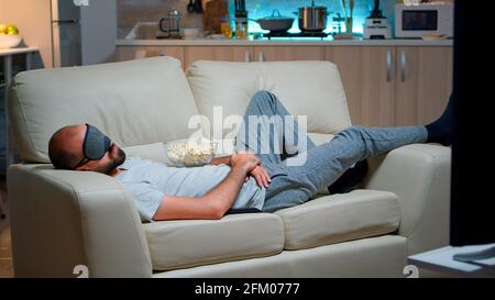 Mann Auge Schlafmaske Einschlafen, während Film, sitzt vor dem Fernseher. Kaukasischer Mann mit Bart aussehende Unterhaltungsserie spät in der Nacht in der Küche Stockfoto