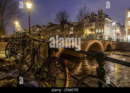 Fahrräder, die in der Winterdämmerung auf der Brücke auf dem eisigen Keizersgracht-Kanal geparkt sind, Amsterdam, Nordholland, Niederlande Stockfoto