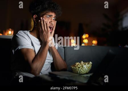 Der junge Mann sieht erschrocken aus, als er alleine mit einem Laptop einen Horrorfilm sieht und Popcorn isst, und sitzt auf einem bequemen Sofa im dunklen Zimmer zu Hause Stockfoto