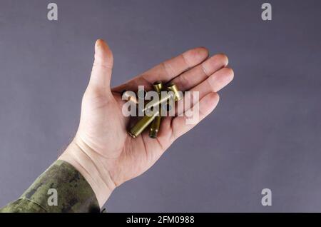 Leere Hüte in der Hand eines Mannes vor grauem Hintergrund. Ein Mann in Tarnuniform hält Pistolen und Maschinengewehr-Gehäuse. Armee-Thema. Milit Stockfoto