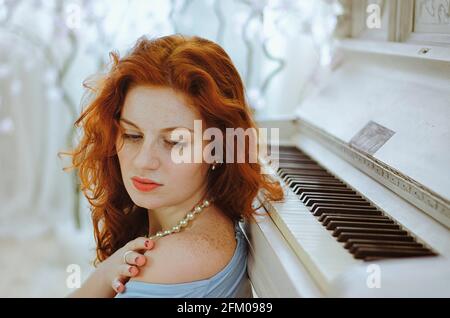 Die wunderschöne rothaarige junge Pianistin mit blauen Augen und Sommersprossen sitzt neben einem Klavier und lernt im Studio Musik. Der kreative Prozess der Herstellung Stockfoto