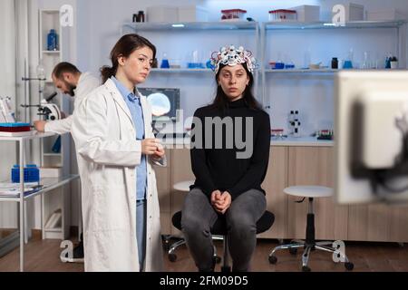 Gestresster Patient, der mit einem eeg-Headset auf einem neurologischen Stuhl sitzt und den Gesundheitszustand auf einem Tablet untersucht. Medizinwissenschaftler, der im Hintergrund durch das Mikroskop schaut und eine Behandlung entwickelt. Stockfoto