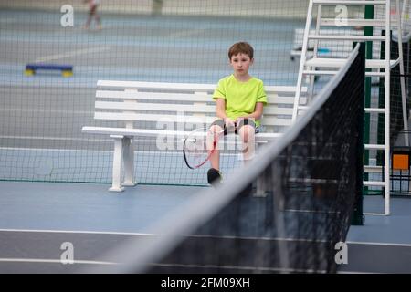 Trauriger Junge sitzt auf einer Bank bei einem Tenniswettbewerb. Das Kind spielt Tennis. Stockfoto