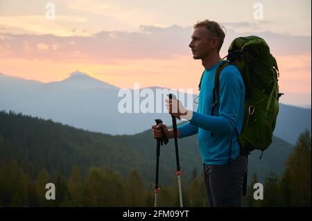 Seitenansicht des männlichen Reisenden, der beim Wandern anhält und in die Ferne schaut und über die nächste Route der Wanderung nachdenkt. Berghügel bedeckt von Wäldern während des Sonnenuntergangs auf dem Hintergrund. Stockfoto
