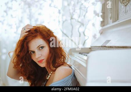 Die wunderschöne rothaarige junge Pianistin mit blauen Augen und Sommersprossen sitzt neben einem Klavier und lernt im Studio Musik. Der kreative Prozess der Herstellung Stockfoto