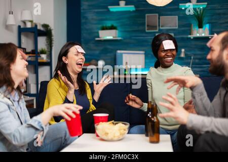 Mixed-Rennen Freunde spielen Brettspiel mit Haftnotizen verbringen Zeit zusammen spät in der Nacht entspannen auf der Couch im Wohnzimmer. Eine Gruppe multiethnischer Menschen, die Bier trinken und Chips essen, genießt die Heimparty. Stockfoto