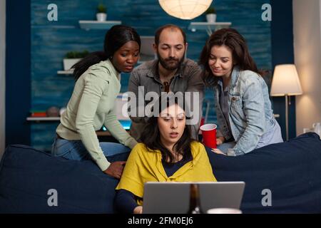 Glückliche Freunde, die sich beim Biertrinken amüsieren und per Videoanruf mit einem Laptop telefonieren. Gruppe von multirassischen Menschen verbringen Zeit zusammen sitzen auf Couch spät in der Nacht im Wohnzimmer. Stockfoto