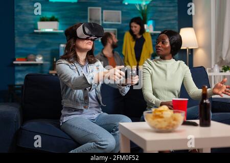 Multiethnische Freunde unterstützen Frau während Videospiel-Wettbewerb spielen mit vr Brille mit Joystick. Gemischte Renngruppe von Menschen, die sich zusammen hingen und sich spät in der Nacht im Wohnzimmer amüsieren. Stockfoto