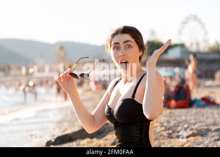 Menschliche Emotionen. Eine Frau sitzt am Strand in einem Badeanzug, ihre Hände sind ungläubig ausgebreitet und bringen Überraschung und Schock zum Ausdruck. Stockfoto