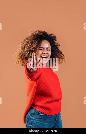 Schöne schwarze Frau mit lockigen Haaren, die Friedenszeichen macht und die Kamera im Studio vor braunem Hintergrund anschaut. Stockfoto
