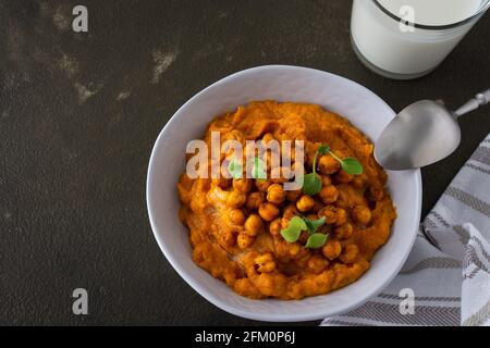 Dicke Kürbissuppe mit gebackenen Kichererbsen und Rettichsprossen auf einem Teller mit einem Glas Milch auf dem Tisch, leckeres gesundes Abendessen Stockfoto