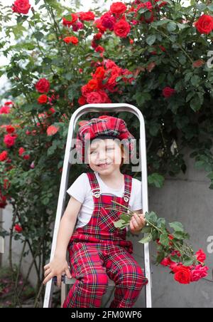 Porträt eines niedlichen fröhlichen Jungen 4 - 5 Jahre alt, in karierten Overalls und sitzender Baskenmütze Auf einer Stube vor dem Hintergrund eines großen Busches bl Stockfoto