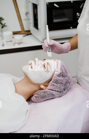 Kosmetologe, der im Spa-Salon eine Maske auf das Gesicht des Kunden anwendet. Junge Frau, die Gesichtsbehandlung durch Kosmetikerin im Spa-Salon Akne-Behandlung, Gesichtspeeling Stockfoto
