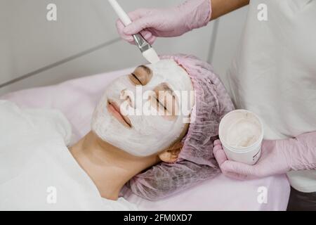Kosmetologe, der im Spa-Salon eine Maske auf das Gesicht des Kunden anwendet. Junge Frau, die Gesichtsbehandlung durch Kosmetikerin im Spa-Salon Akne-Behandlung, Gesichtspeeling Stockfoto