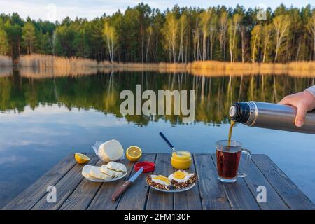 Im Frühling wird aus nächster Nähe von einer Thermoskanne am Morgen neben dem See und dem Wald heißer Tee in einen Glaskrug gegossen. Frühstück auf einem Holztisch. Natu Stockfoto