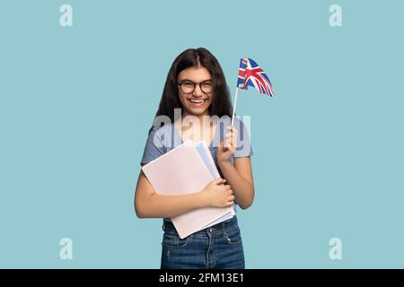 Positiver indischer Teenager-Austauschstudent mit Notizbüchern mit britischer Flagge, Blick auf die Kamera und einem Lächeln auf blauem Hintergrund Stockfoto