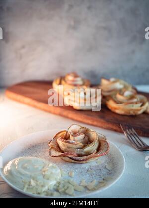 Apfelrosen, Apfelbrötchen, serviert mit Schlagsahne und Mandelblüten, grauer Hintergrund. Stockfoto