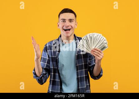 Hübscher junger Mann, der sich über den Gewinn einer großen Geldsumme freut und Dollarscheine auf orangefarbenem Studiohintergrund hält Stockfoto