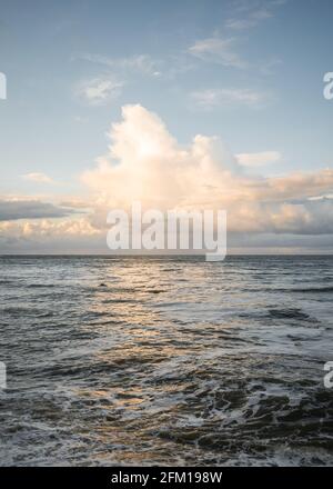 Wunderschönes goldenes Licht, das von Wolken über dem Meer reflektiert wird und Ozeanwellen am Horizont und Wasser mit cremig-goldenen, ruhigen Farben an der Küste erhellt Stockfoto