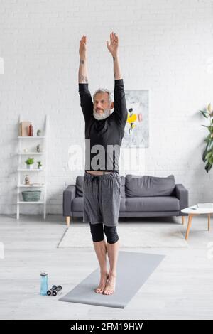 Die ganze Länge der bärtigen und tätowierten Mann trainieren auf Fitness Matte neben Sportflasche und Hanteln im Wohnzimmer Stockfoto