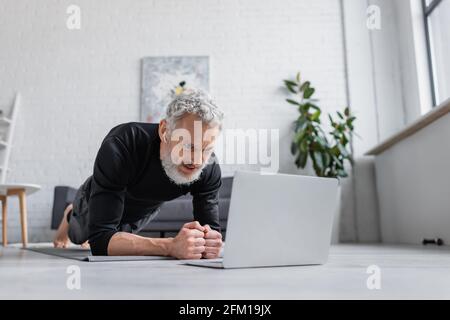 Bärtiger Mann in kabellosen Kopfhörern, der auf der Fitnessmatte eine Planke machte In der Nähe des Laptops im Wohnzimmer Stockfoto