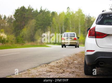 Weißer SUV, der am Straßenrand in einem wunderschönen Frühlingswald geparkt ist. Eine asphaltierte Straße zwischen grünen Bäumen an einem Frühlingstag. Mit dem Auto durch ein einheimischer Coun Stockfoto