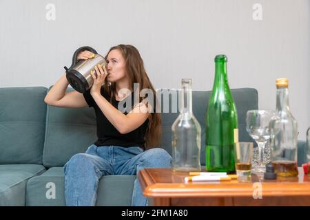 Die junge dunkelhaarige Frau sitzt mit offenen Augen auf einem Sofa und trinkt nach einer wilden Party Wasser aus einer Teekannen, neben ihr stehen leere Flaschen Stockfoto