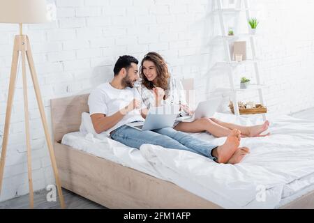 Fröhlicher, multiethnischer Freiberufler, der auf dem Bett liegt und Laptops benutzt Stockfoto