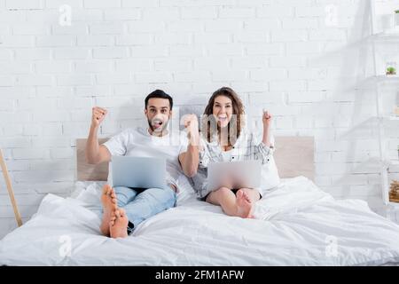 Glückliche, multiethnische Freiberufler, die mit Laptops auf dem Bett liegen Stockfoto