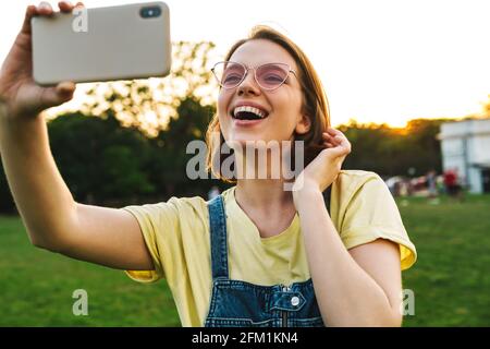 Bild der fröhlichen Brünette Frau, die Selfie-Foto auf dem Handy Und lächeln, während Sie sich im Sommerpark ausruhen Stockfoto